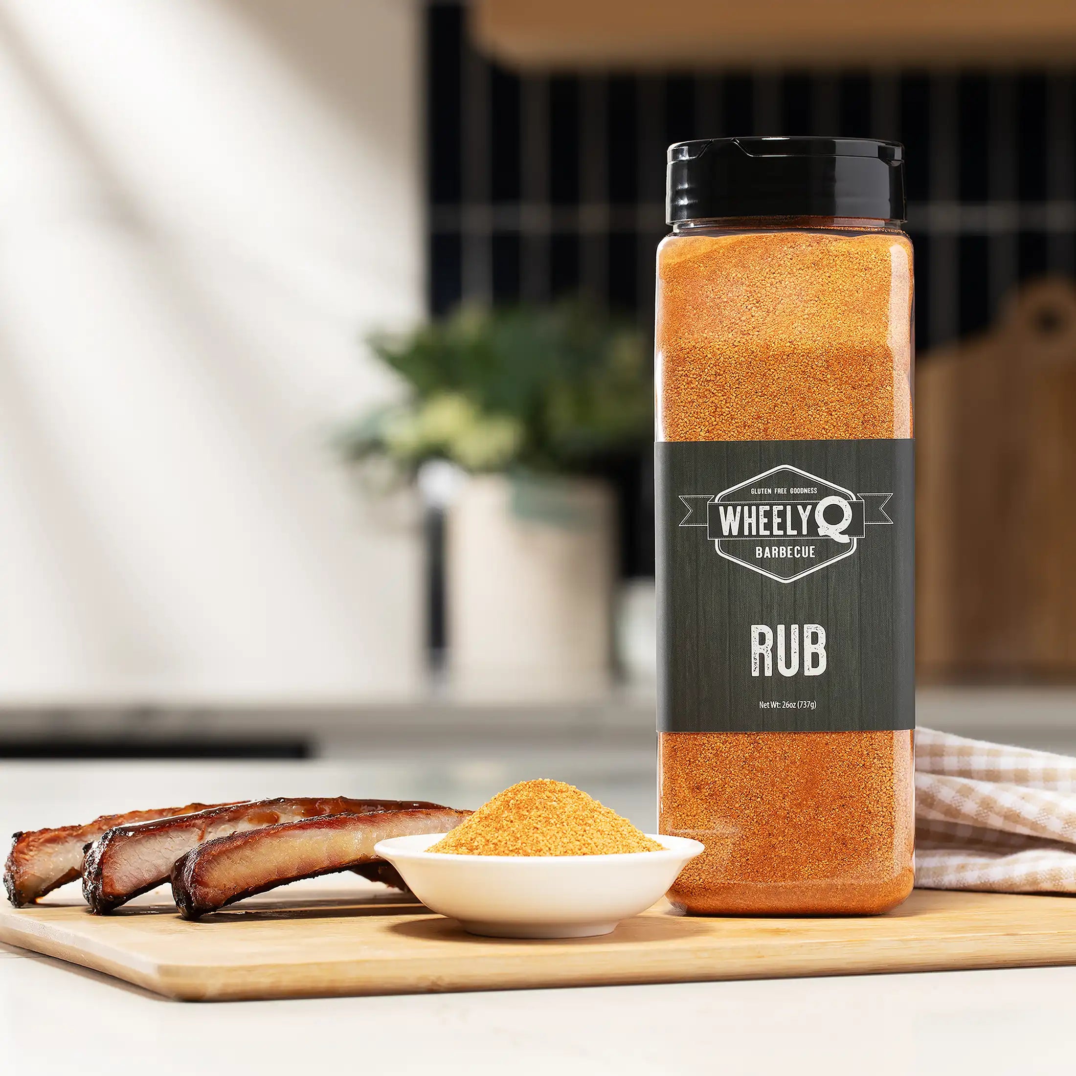 WheelyQ Barbecue All-Purpose Rub 26oz shaker bottle sitting on a cutting board in a kitchen.  On the cutting board is also a little white bowl full of the seasoning with 3 ribs on the left.