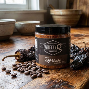 Jar of WheelyQ Barbecue Ancho Espresso Seasoning on a wooden surface with coffee beans and chilis.