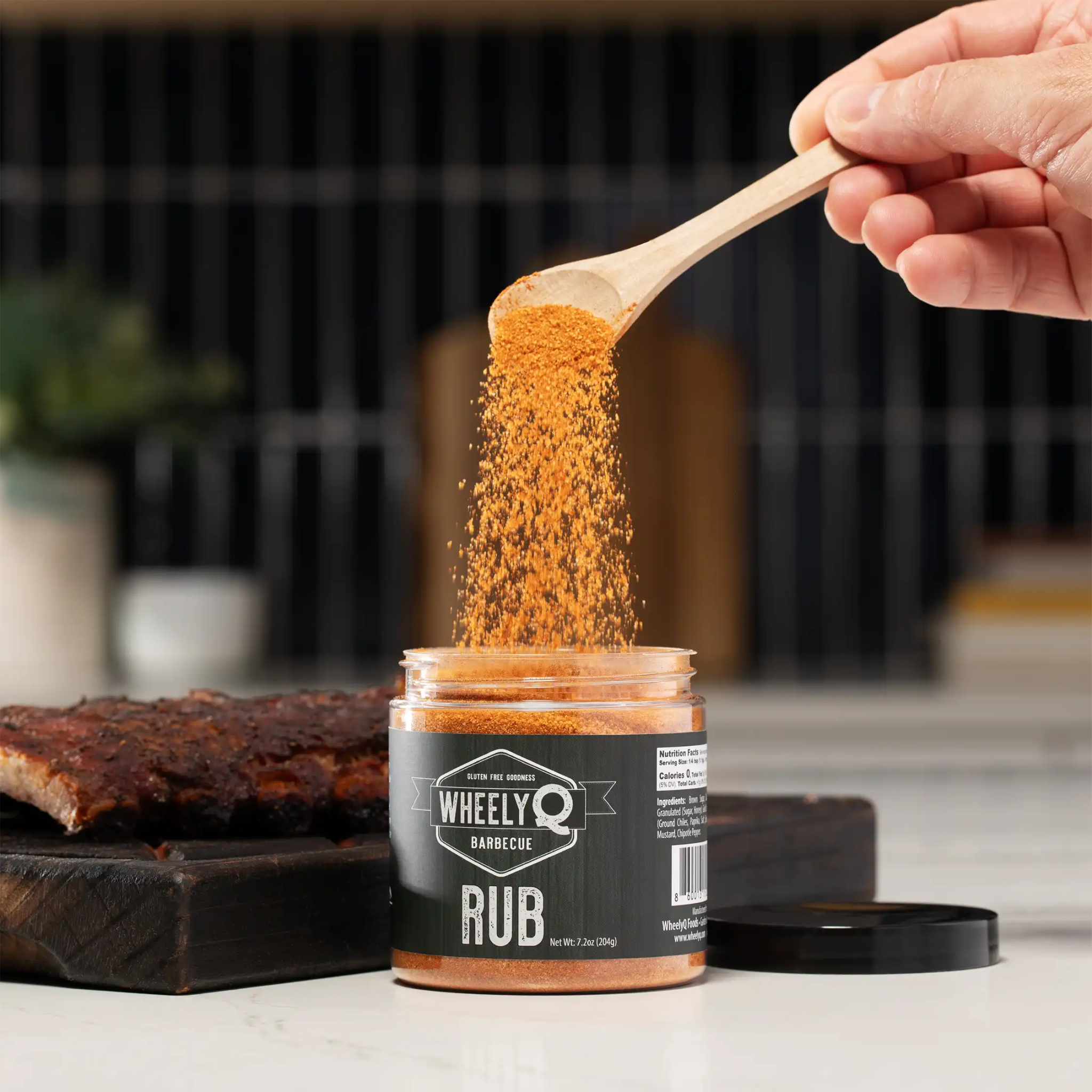 WheelyQ Barbecue All-Purpose Barbecue Rub being poured from a wooden spoon into a jar with a blurred kitchen background.