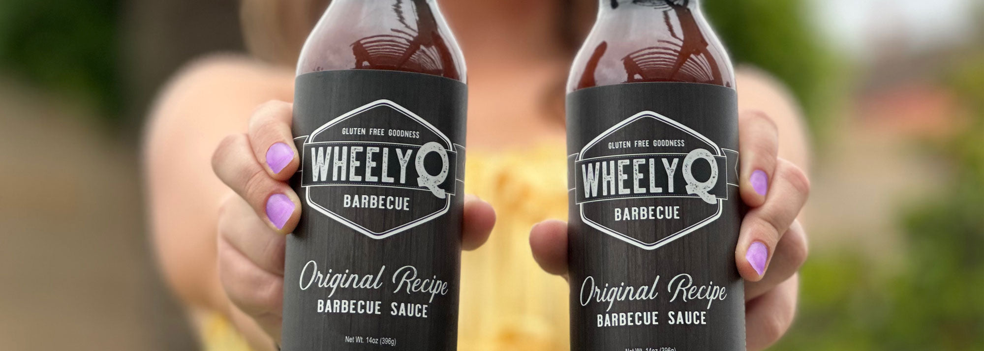 Image of two bottles of WheelyQ Barbecue Sauce being held by a beautiful girl that is faded in the background.