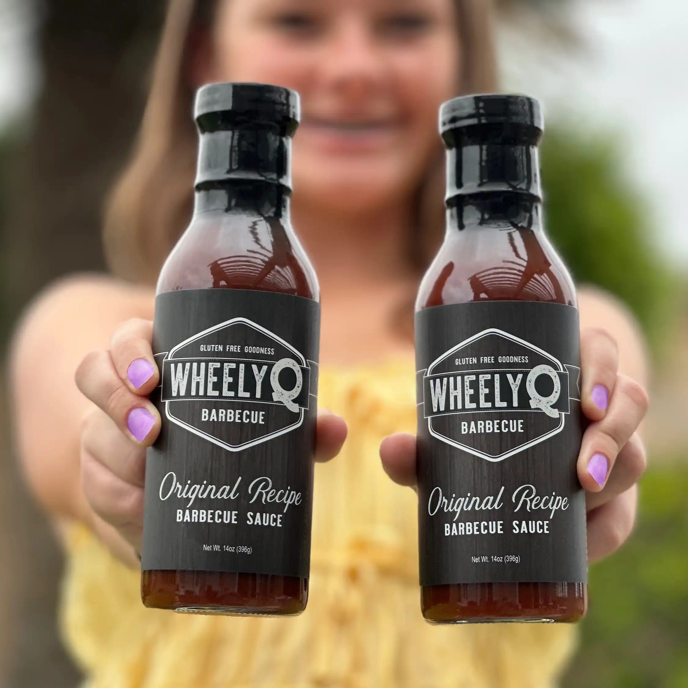 Image of two bottles of WheelyQ Barbecue Sauce being held by a beautiful girl that is faded in the background.