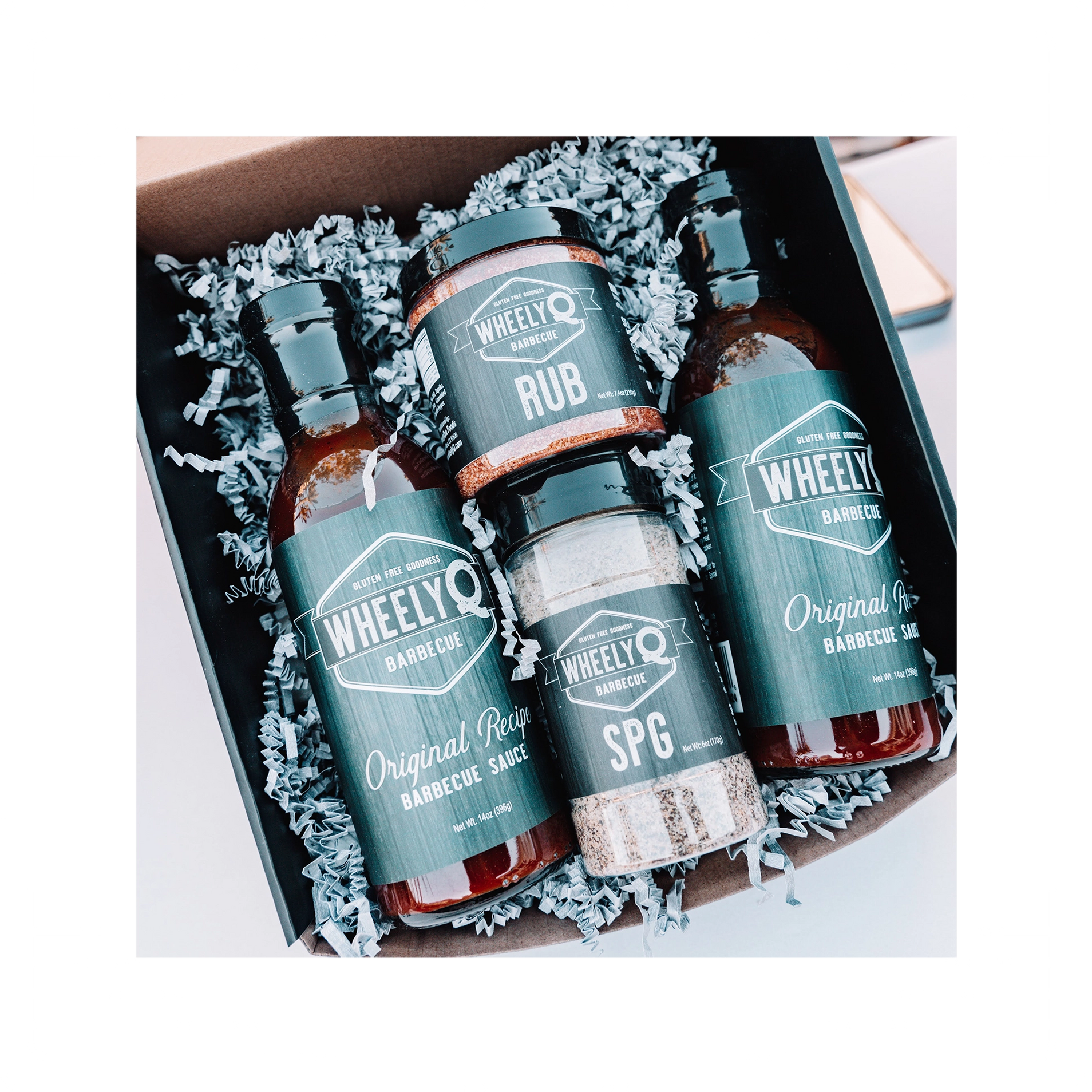 An image of a Barbecue Gift Box sold by WheelyQ Barbecue.  The gift set include 2 bottles of Barbecue Sauce, 1 bottle of SPG Seasoning and 1 jar of All-Purpose Rub or Ancho Espresso Seasoning.