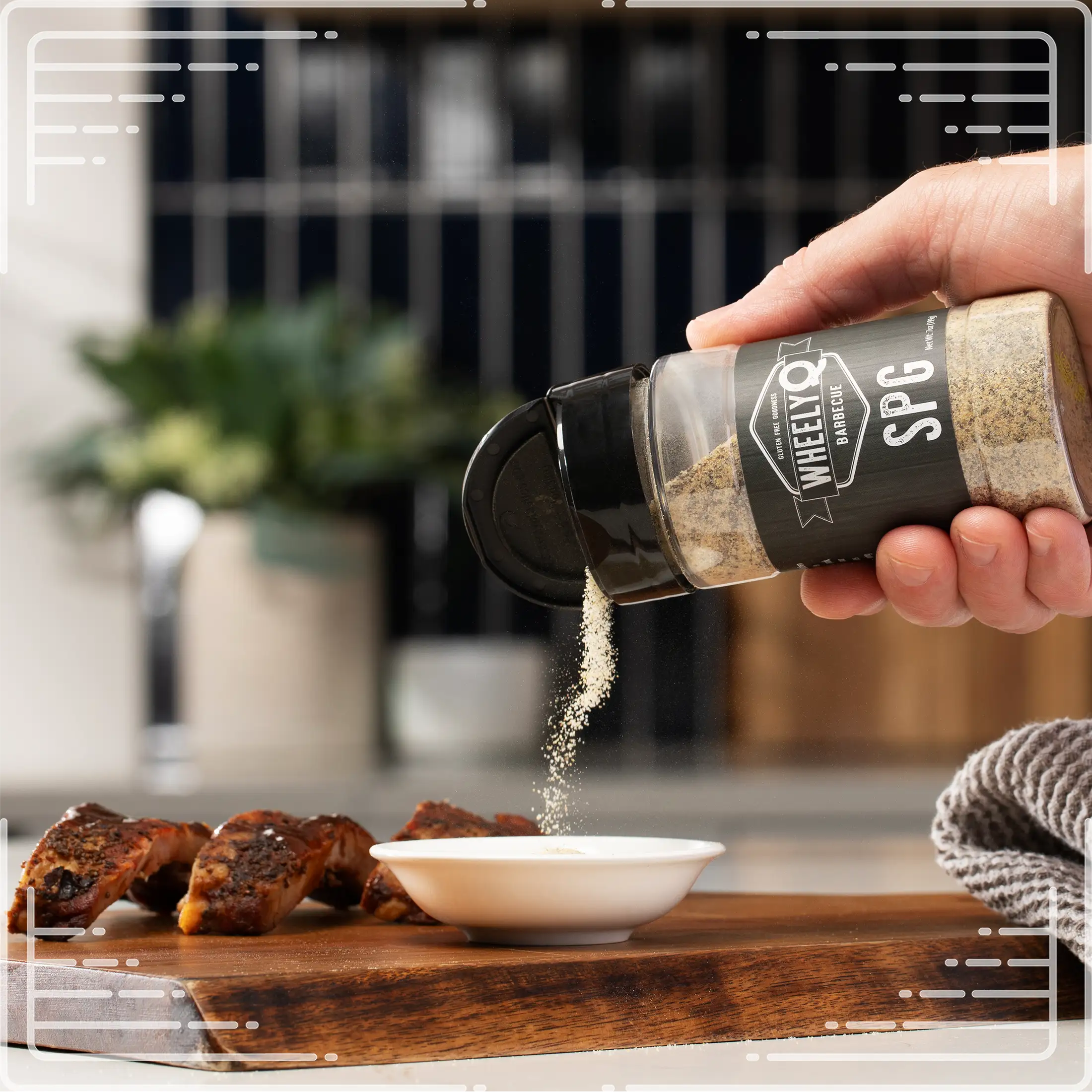 Hand sprinkling WheelyQ SPG Seasoning (Salt, Pepper, Garlic) from a spice jar into a small white bowl n a wooden cutting board.