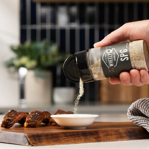 Hand sprinkling WheelyQ SPG Seasoning (Salt, Pepper, Garlic) from spice jar into a small white bowl on wooden cutting board.